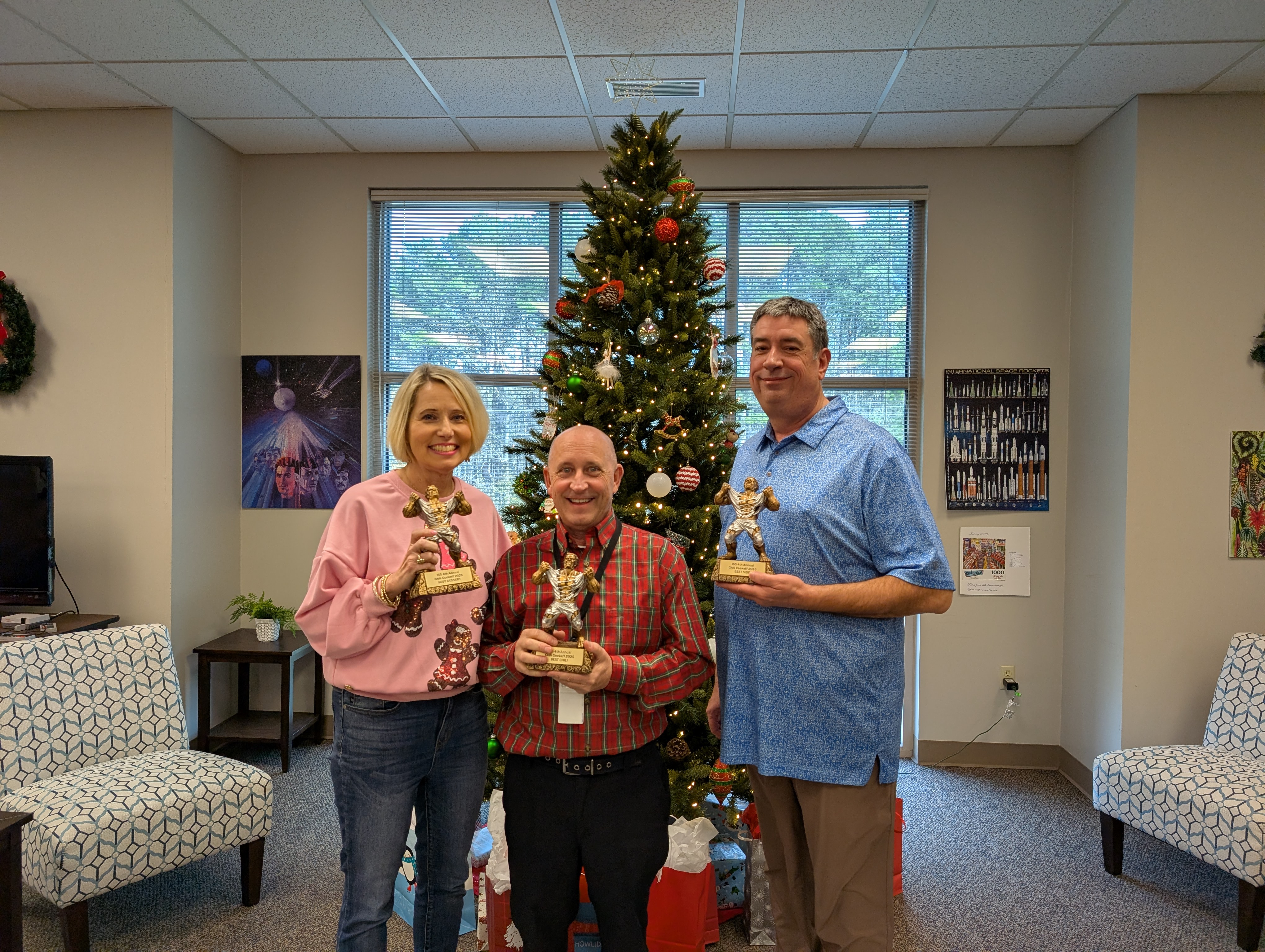 Michelle, Greg, and Devin standing in front of the Christmas tree with their trophies for winning the Chili Cookoff for 2025